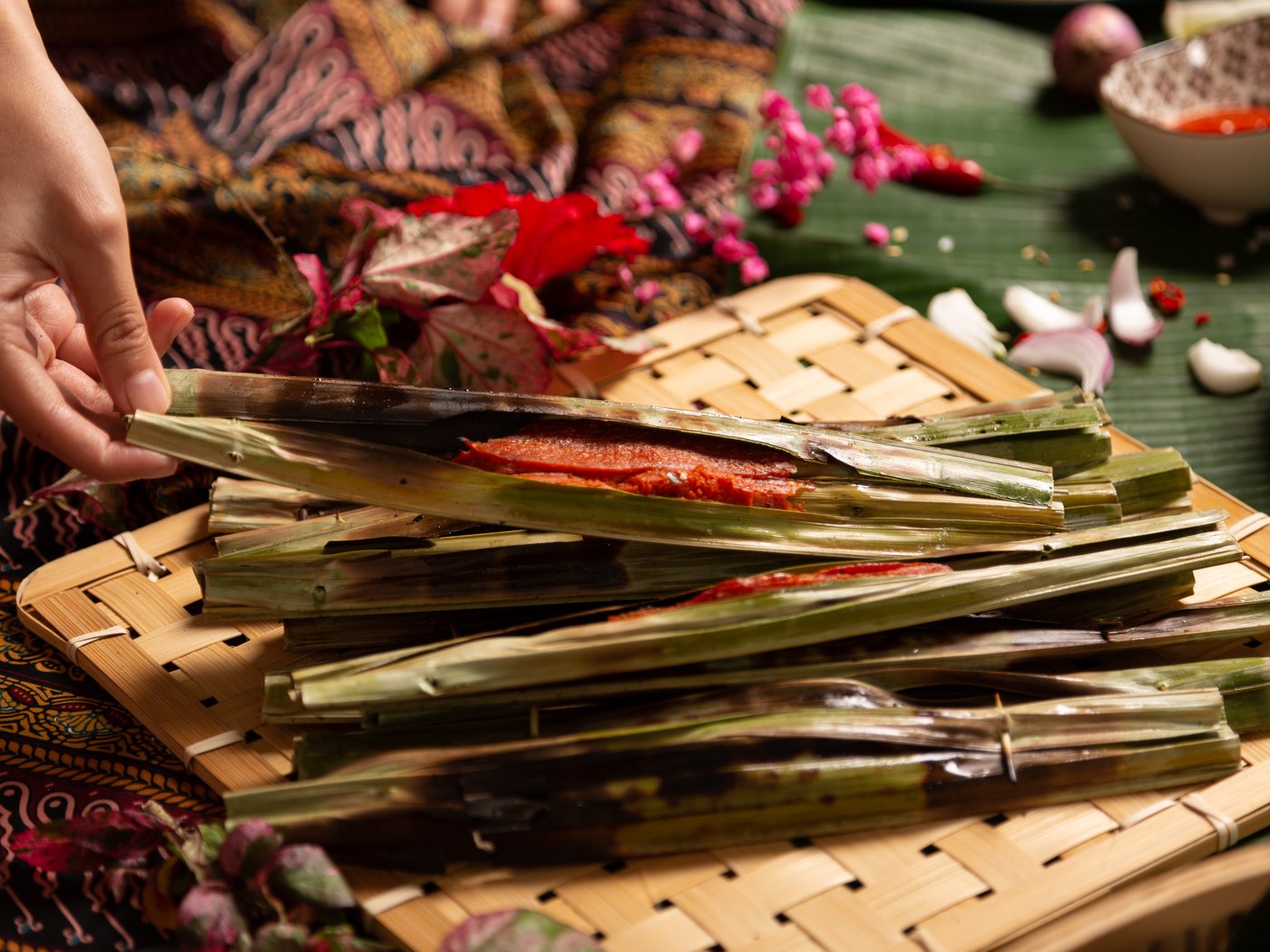A hand lifting an otak-otak parcel from a woven tray